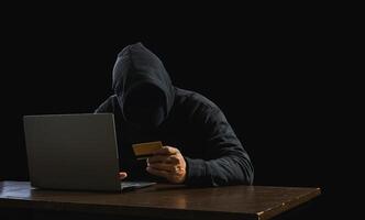 Portrait hacker spy man one person in black hoodie sitting on table looking computer laptop used login password attack security to data digital internet network system night dark background copy space photo