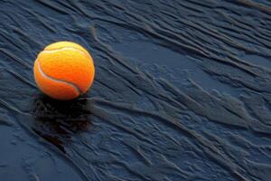 Bright orange tennis ball resting on a textured dark surface in a dramatic setting photo