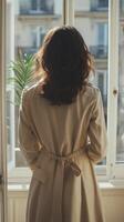 Woman in beige coat observing city view through window in bright room photo
