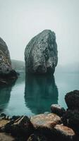 Mysterious rock formation rises from calm water on foggy morning near coastal cliffs photo