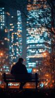City dweller working on laptop at night in urban park with glowing skyscrapers photo
