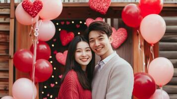 A couple poses happily in front of a festive backdrop decorated with red and pink hearts, balloons, and twinkling lights. photo