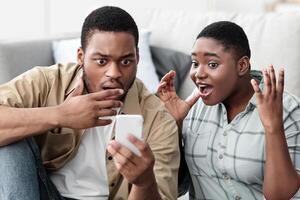 Shocked Black Tourists Couple Using Cellphone Reading Shocking Travel News Or Negative Message About Tourism Sitting Near Suitcase At Home. Oh No, Flight Is Cancelled. Traveling Problems Concept photo