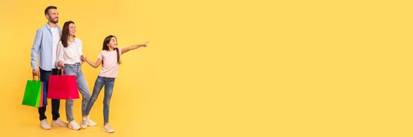 Family Shopping. Happy Parents And Little Daughter Holding Shopper Bags, Pointing Finger Aside At Copy Space, Asking Mom And Dad To Buy Something, Showing Great Offer Over Yellow Backdrop, Panorama photo