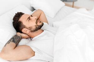 Thoughts in head not let fall asleep. Millennial man lying in bed and looking at ceiling, thinking of something in the morning, free space photo