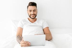 Portrait of happy man sitting on bed under blanket and using computer, typing on laptop keyboard, browsing internet, working or resting, free space photo