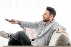 Relaxed man switching channels with remote controller while watching tv at home, relaxing on couch in living room, enjoying weekend pastime, copy space, side view photo