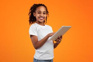Digital Natives, App For Kids. Beautiful smiling African American teenage girl holding and using digital tablet over orange studio background, looking and posing at camera, banner with copy space photo