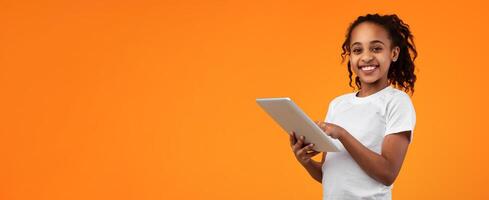 Digital Natives, App For Kids. Beautiful smiling African American teenage girl holding and using digital tablet over orange studio background, looking and posing at camera, banner with copy space photo