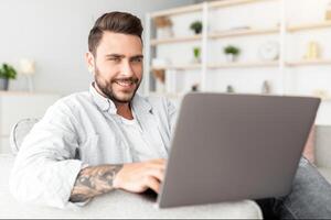 Positive guy reclining on sofa with laptop at home interior, panorama, copy space. Handsome man using notebook, watching movie, surfing internet or scrolling social networks photo
