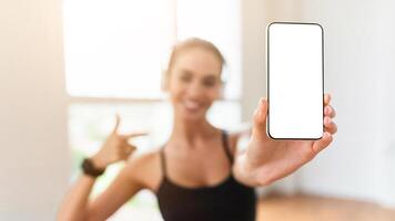 Sports App Concept. Fit young lady holding and showing smartphone with white empty screen for mock up template, pointing at isolated gadget display, blurred background, selective focus on hand photo