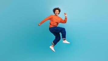 A young African American man with curly hair is jumping joyfully against a vibrant blue background. He is wearing a casual orange sweater and blue jeans, exuding youthful energy and enthusiasm. photo