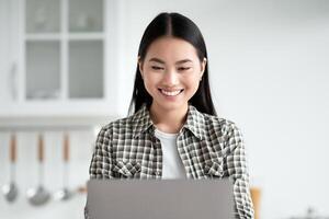 Joyful asian lady sitting at kicthen, working online, using modern laptop, typing on keyboard, sending emails or chatting with clients. Young woman student doing homework, copy space photo