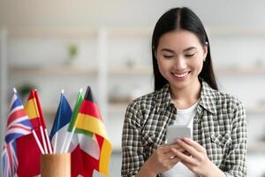 Cheerful asian young woman using cellphone, having English or other foreign language class, sitting at table with wireless headset and international flags at home, using mobile application, copy space photo