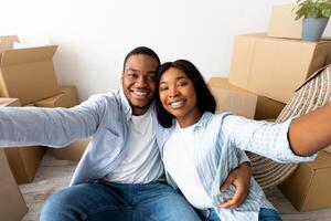 Excited african american couple making selfie after moving to own house. Spouses sitting near unpacked boxes and smiling at home. Relocation, house purchase and ownership concept photo