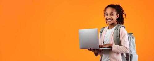 Portrait of smiling African American girl holding and using laptop computer, wearing backpack, typing on keyboard, posing and looking at camera standing isolated over yellow studio background photo