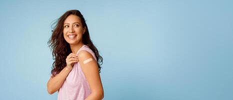 Vaccination Saves Lives. Joyful Arab Woman Showing Vaccinated Arm With Medical Bandage After Coronavirus Antiviral Vaccine Injection Posing On Blue Background. Stop Coronavirus. Panorama, Copy Space photo
