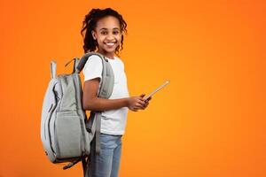 Modern Education And Technology. Portrait of smiling black girl holding and using digital tablet, wearing backpack,posing and looking at camera standing isolated on yellow studio wall, free copy space photo