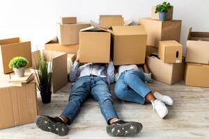Moving day. Tired african american couple with cardboard boxes on heads, sitting on floor, being tired in unpacking things while relocating to new apartment photo