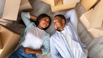 Loving black family with carton boxes around, lying on floor and having rest on moving day, top view. Black woman with her husband looking at each other and smiling, overhead view photo