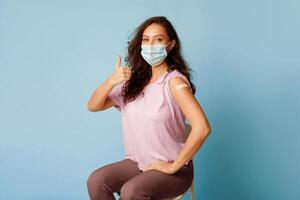 Covid-19 Vaccination. Vaccinated Woman Gesturing Thumbs Up Showing Arm With Plaster Bandage After Coronavirus Vaccine Injection On Blue Background, Wearing Protective Face Mask. Antivital Immunization photo
