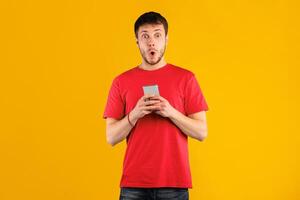 Wow, Amazing Application. Portrait of excited surprised young man using cellphone standing isolated over blue studio background, copy space. Socked guy chatting online, looking at camera photo