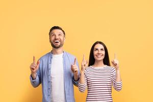 A happy couple stands side by side, pointing excitedly upwards. They share smiles and look thrilled, showcasing their affection. The bright yellow backdrop adds a lively touch to the moment. photo