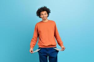A young African American man expresses a playful demeanor with empty pockets, highlighting a common challenge faced by millennials in today economy while posing against a bright blue backdrop. photo