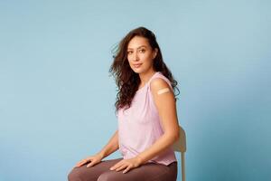 Stop Coronavirus, Vaccination Campaign. Vaccinated Woman Showing Arm Sitting With Rolled Up Sleeves Posing After Covid-19 Vaccine Injection On Blue Background. Studio Shot photo