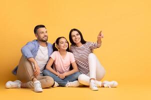 A loving family of three shares a joyful moment against a bright yellow background. The parents sit closely with their daughter, who points at copy space while they all smile photo