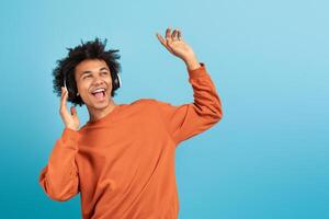 A young African American man in a bright orange sweater dances with headphones on, completely immersed in the music. He has a joyful expression against a blue background, copy space photo