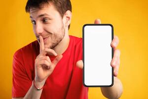 Man with white blank cellphone screen holding index finger on lips, showing gesture of secret, empty space, mock up photo