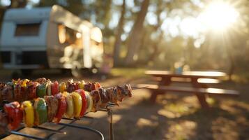 Skewers of colorful grilled kebabs sizzle over a fire as the sun sets in a serene camping spot. A caravan and picnic table complete the picturesque outdoor setting. photo