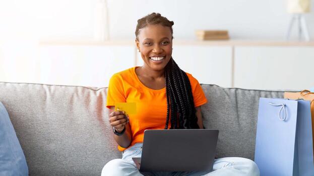 hermosa afro dama compras en línea, sentado en sofá con ordenador portátil ordenador personal, participación crédito tarjeta, regalo pantalones en pie cercano. encantador milenario negro mujer adquisitivo regalos en web desde hogar foto
