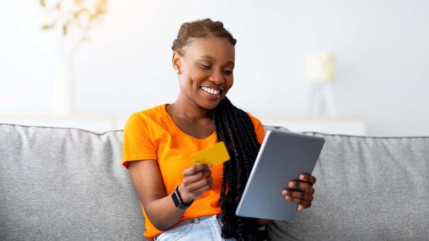 alegre negro mujer haciendo en línea pago, comprando bienes en web, utilizando Internet banco servicios, sentado en sofá a hogar con tableta ordenador personal y crédito tarjeta. Internet compras, estacional venta, negro viernes foto