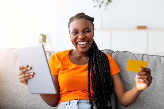 comercio electrónico y en línea compras concepto. retrato de positivo africano americano mujer compras en Internet, utilizando digital tableta y crédito tarjeta, sentado en sofá a hogar. ventas y descuentos foto