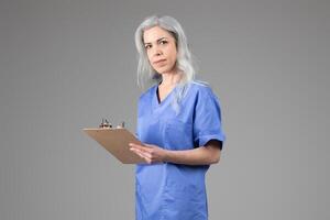 A middle aged woman dressed in scrubs holds a clipboard while looking at the camera. Her silver hair contrasts with her blue uniform, emphasizing her role in healthcare during a calm moment. photo