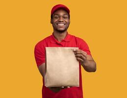 Fast Delivery. Front view of smiling african american courier guy in red cap and uniform holding and giving craft paper bag from restaurant with products to camera, yellow orange studio background photo