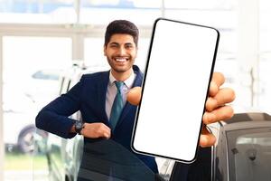 Emotional middle eastern businessman choosing new car, standing by brand new black auto, showing mobile phone with white blank screen and smiling, using auto dealership app, mockup photo