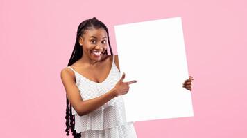 Smiling African American woman pointing at empty banner with space for your design on pink background. Attractive black lady with afro bunches showing blank poster, mockup for summer ad photo