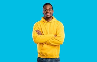 Handsome Black Millennial Man Crossing Hands Posing Smiling To Camera Standing In Studio On Blue Background, Wearing Yellow Hoodie. Shot Of Confident African American Guy photo