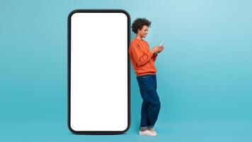 A young African American man stands next to an oversized smartphone, engaging with his device. He appears joyful, showcasing a casual lifestyle in a vibrant blue setting. photo