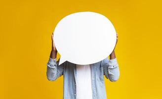 Unrecognizable black guy holding empty speech bubble in front of his head over yellow studio background, copy space photo
