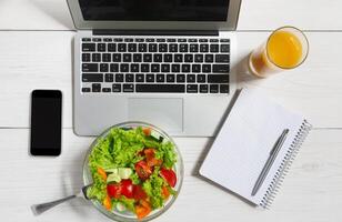 Healthy business lunch in the office, top view of vegetable salad on white wooden desk near laptop computer keyboard. Salad bowl, juice, mobile phone and notepad with pen flat lay. Snack at break time photo