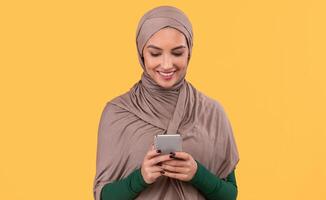 Cheerful Arabic Lady Using Mobile Phone Texting Online And Networking In Social Media Standing Over Yellow Background In Studio. Happy Smartphone User, Cellphone Application Advertisement Concept photo