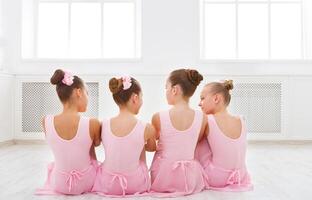 Little ballerinas talking in ballet studio. Group of girls having break in practice, sitting on floor, back view. Classical dance school photo