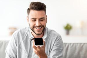 Happy handsome man recording voice message on smartphone or using virtual assistant while resting on sofa at home. Smiling guy enjoying smart technologies for communication, free space photo
