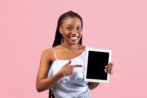 Positive young Afro woman pointing at tablet computer with empty screen on pink studio background, mockup for website or app. Cool black lady presenting touch pad with space for ad photo