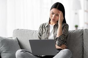 Exhausted korean lady suffering from headache while using laptop, sitting on couch and working from home, touching her head, copy space. Tired young asian woman having problems with computer photo