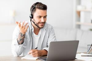 Web conference. Young freelancer man in headset making call on laptop, sitting at workplace at home, talking and gesturing at webcamera, free space photo